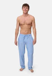 Kaal bovenlijfs jonge man die op blote voeten staat en lichtblauwe joggingbroek met koord draagt, op een effen lichte achtergrond.