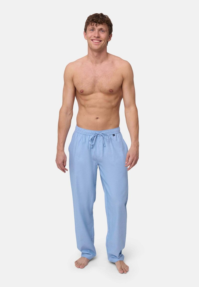 Kaal bovenlijfs jonge man die op blote voeten staat en lichtblauwe joggingbroek met koord draagt, op een effen lichte achtergrond.