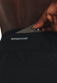 Shorts deportivos negros con una cinturilla ajustada y una apertura para un bolsillo marcado como "SPEEDPOCKET." Una mano está insertando un dispositivo en el bolsillo.