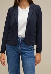 Námořnický blazer s hladkou texturou, dvěma předními kapsami a zapínáním na jeden knoflík. Má klopový límec a žebrované manžety. Nosený přes bílou košili.