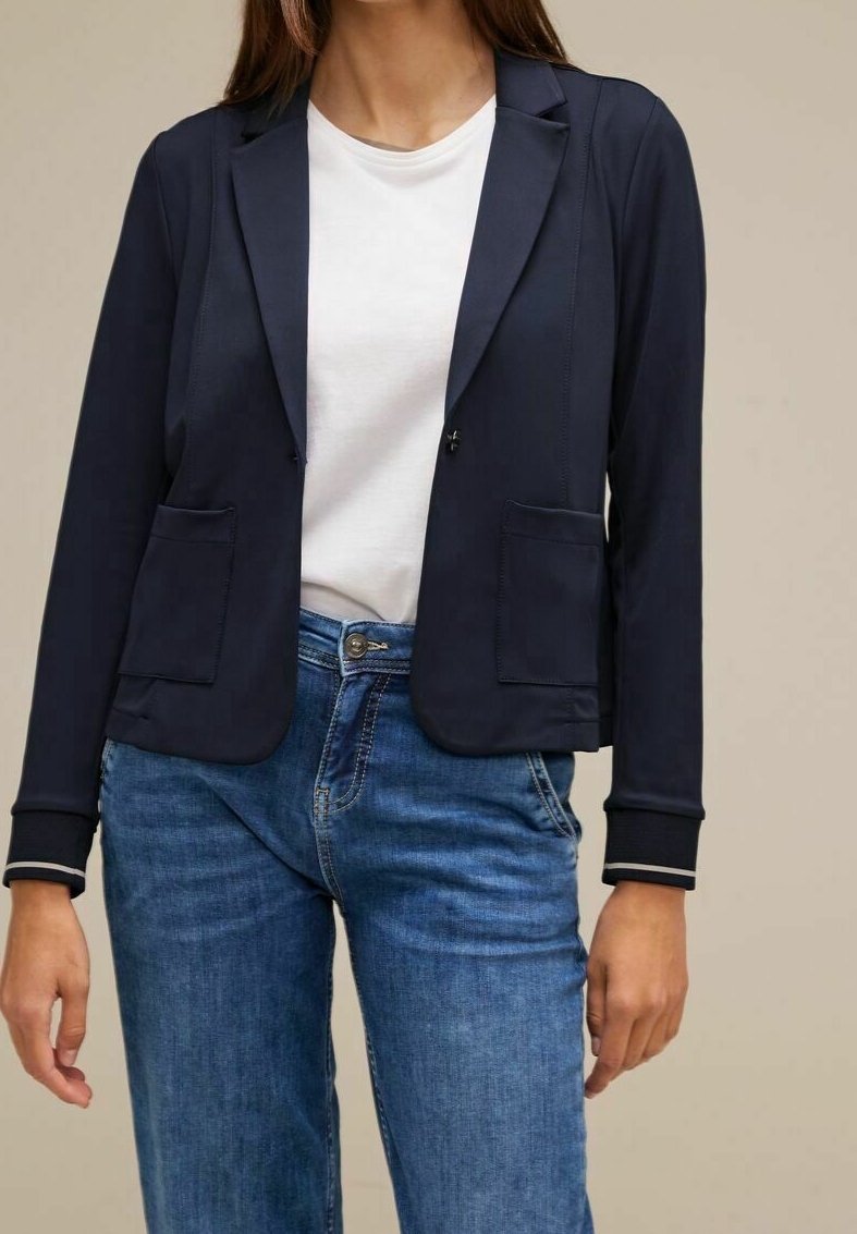 Námořnický blazer s hladkou texturou, dvěma předními kapsami a zapínáním na jeden knoflík. Má klopový límec a žebrované manžety. Nosený přes bílou košili.