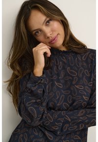 Marinebluse med paisley-mønster i kobbertoner, med rynket halsudskæring og knaplukning ved manchetterne. Blødt stof med tekstureret finish.