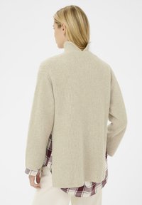 Maglione a coste beige con collo alto e spacchi laterali, indossato sopra una camicia a quadri bordeaux e bianca. Texture morbida, vestibilità rilassata.