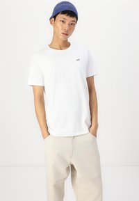 White cotton t-shirt worn with beige pants. T-shirt features a small black logo on the chest. Model has a blue striped beanie on.