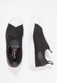 Černé nazouvací tenisky s bílou gumovou špičkou, elastickými křížovými pásky a texturovaným látkovým svrškem. Na patě mají poutko pro snadné nazouvání.