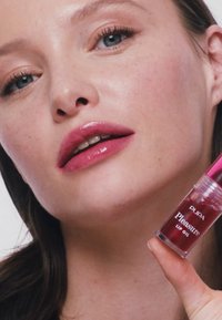 Olio per labbra trasparente in un contenitore trasparente con accenti rosa. Il modello ha un aspetto naturale, mostrando labbra rosa lucide e pelle liscia.