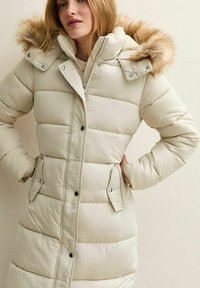 Mujer con un abrigo de plumas largo beige, capucha con borde de piel, botones frontales y bolsillos con solapa, de pie con las manos en las caderas.