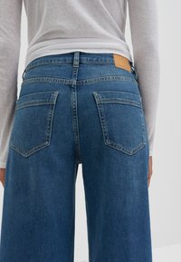 Jeans en denim bleu à jambes larges avec un lavage clair, comportant deux poches arrière à patch et une pièce rectangulaire en cuir tan sur la ceinture.