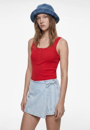 Ung kvinde med langt hår iført en blå bucket hat, rød ærmeløs top med knapper og en lyseblå denim omslagsnederdel med sidebinding.