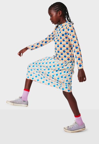 Langärmliges Kleid mit weißem Hintergrund, das bunte geometrische Muster in Blau, Orange und Gelb zeigt. Kombiniert mit grauen Turnschuhen.