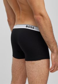 Boxeurs noirs avec une ceinture élastique blanche portant le logo « BOSS ». Tissu lisse avec un design ajusté, laissant voir la peau en dessous de la ceinture.