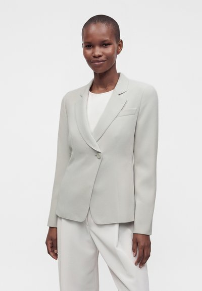 Personne portant un blazer gris clair sur mesure sur un haut blanc et un pantalon large assorti, debout devant un fond blanc uni.
