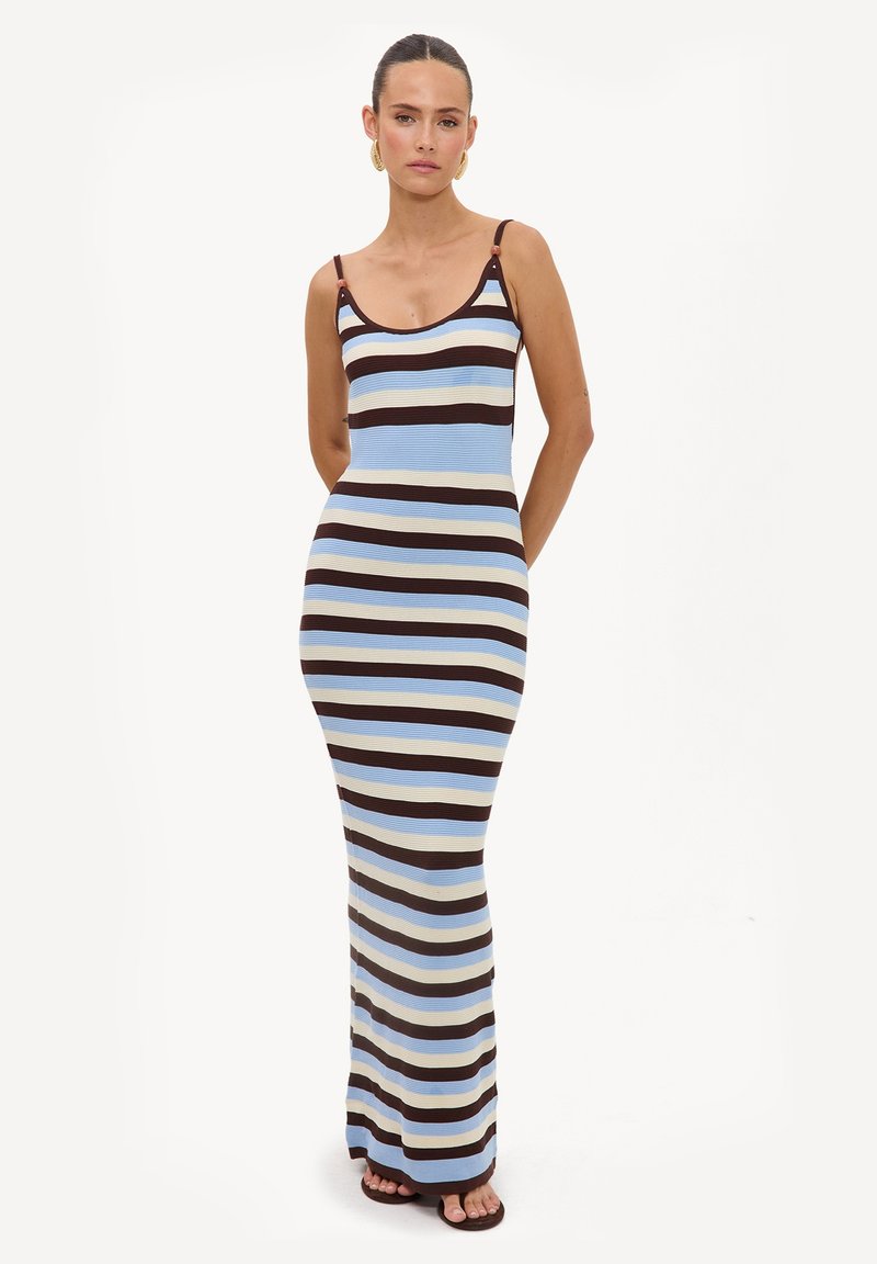 Femme portant une robe sans manches, longue jusqu'au sol, rayée en bleu, brun et beige, les mains dans le dos et expression neutre.
