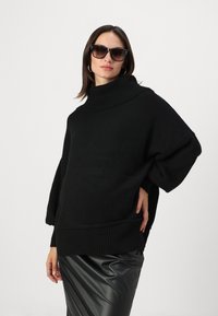 Pull en tricot surdimensionné noir avec un col côtelé haut et des manches larges. Associé à une jupe en cuir noir brillant.