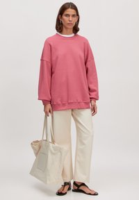 Overmaatse roze sweatshirt, crèmekleurige wijde broek, zwarte platte sandalen en een beige tote bag met een patchzak. Eenvoudig, ontspannen ontwerp.