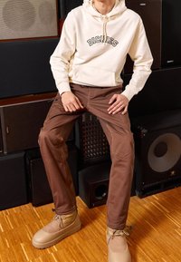 Sudadera con capucha beige con el logotipo "DICKIES", combinada con pantalones ajustados marrones y botas de tobillo beige, todo esto frente a un equipo de audio oscuro.