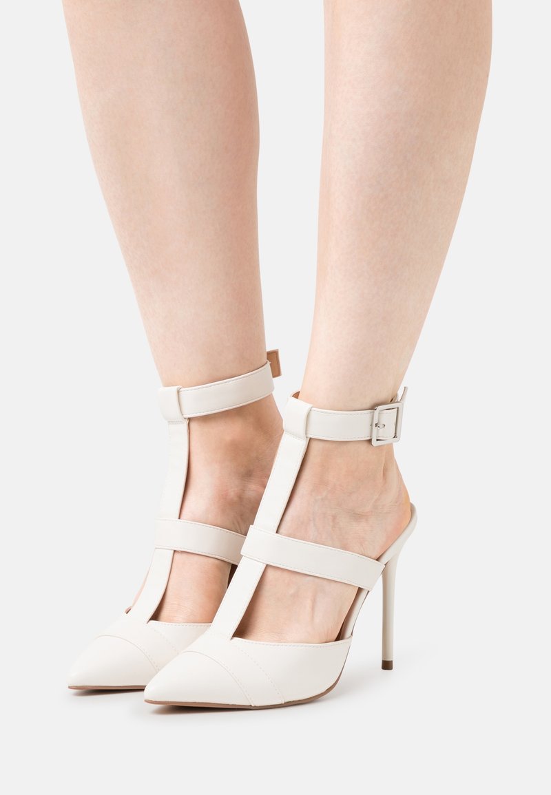 Pieds portant des escarpins blancs à bout pointu avec des brides à la cheville et une sangle verticale reliant les orteils à la cheville sur un fond clair.