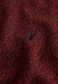 Red and black textured knit fabric with a small black embroidered stag logo near the center.