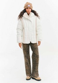 Biała kurtka puchowa z wysokim kołnierzem i beżowymi paskami jako akcentem, zestawiona z brązowymi spodniami w panterkę i beżowym obuwiem.