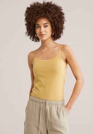 Žena s kučeravými vlasmi, ktorá nosí zlaté kruhové náušnice, horčicovo žlté tielko a béžové nohavice so šnúrkou, stojaca pred jednoliatym pozadím.