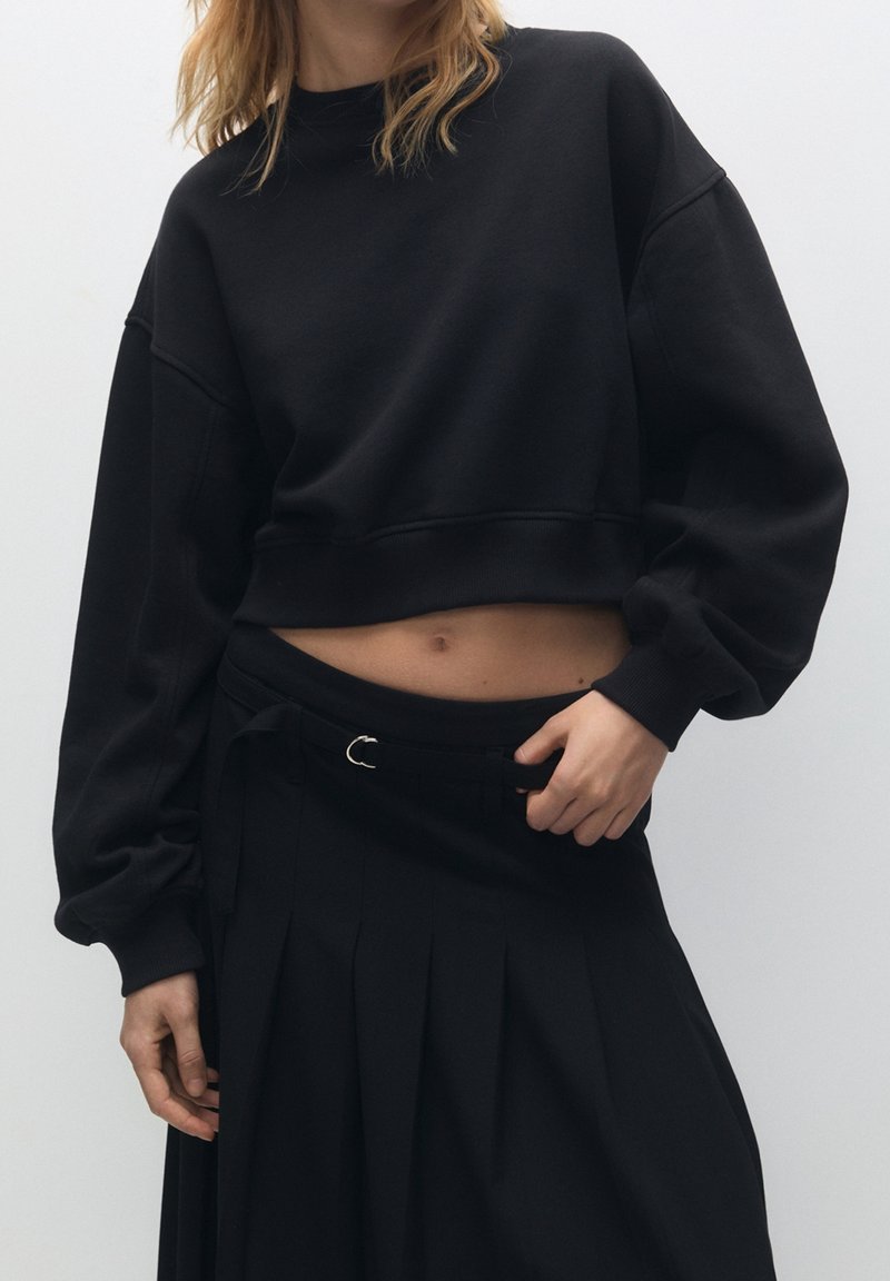 Femme portant un sweat court noir et une jupe noire plissée taille haute avec ceinture, debout devant un fond clair uni.