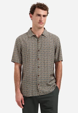 Mann trägt kurzärmeliges Hemd mit braunem, blauem und beigem geometrischem Muster, steht mit einer Hand in der Tasche vor weißem Hintergrund.