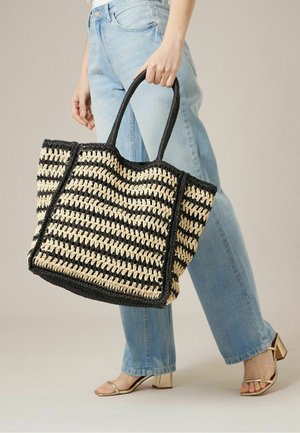 Main tenant un grand sac fourre-tout tissé rayé noir et beige, portant un jean bleu clair et des sandales à talons blocs ouvertes beige.