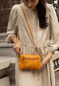 Kvinna i beige klänning som håller en liten senapsgul axelväska med framficka, stående nära stensteg och tegelvägg.