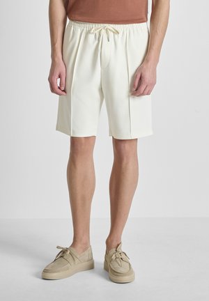 Uomo che indossa pantaloncini bianchi sporchi fino al ginocchio con coulisse e scarpe beige con lacci, in piedi su uno sfondo chiaro uniforme.