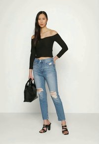 Top preto off-the-shoulder, jeans claros rasgados, sandálias pretas de salto alto e uma pequena bolsa preta. Modelo está de pé contra um fundo neutro.
