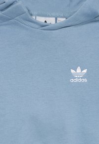 Ljusblå hoodie med vit Adidas Trefoil-logotyp broderad på vänster bröstområde.