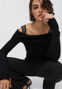 Črni rebrasti top s spuščeni rameni in naramnicami z zakovicami, dolgimi rokavi ter telesno prilegajočo silhueto, kombiniran s spletnimi črnimi hlačami in visokimi petami.