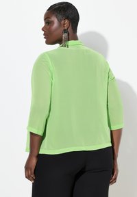 Blusa trasparente di colore verde neon con maniche 3/4, orlo dritto e vestibilità rilassata. Indossata sopra pantaloni neri, aggiunge una sottile texture e stratificazione.