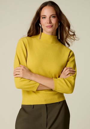 Kvinde med langt brunt hår iført gul sweater og grønne bukser, står med armene over kors og neutral ansigtsudtryk på beige baggrund.