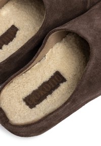Een paar bruine suède pantoffels met een zachte, roomkleurige fleecevoering en een donkerbruine merketiket aan de binnenkant.