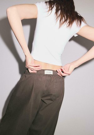 Person trägt ein weißes Kurzarmhemd und lockere braune Hosen, die Hände justieren die Hosen am unteren Rücken, vor schlichtem hellem Hintergrund.