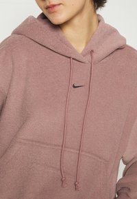 Sweat à capuche en polaire rose avec une finition texturée, des cordons de serrage réglables, une poche avant et un petit logo Nike noir sur la poitrine.