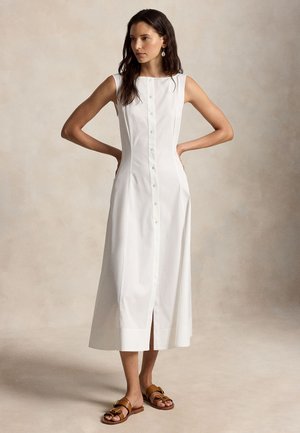 Γυναίκα με μακριά σκούρα μαλλιά, φορώντας λευκό midi φόρεμα χωρίς μανίκια με κουμπιά μπροστά και καφέ σανδάλια με αγκράφα, στέκεται με τα χέρια στη μέση.