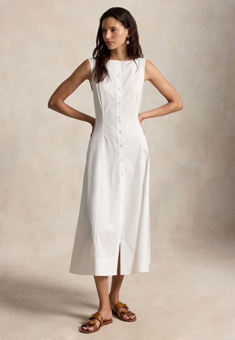 Woman with long dark hair wearing sleeveless white button-front midi dress and brown buckle sandals, standing with hands on hips.