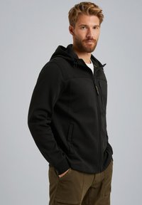 Sudadera negra con cremallera y capucha, que cuenta con un bolsillo frontal, bolsillos laterales y una textura suave. Combinada con pantalones cargo verde oliva.