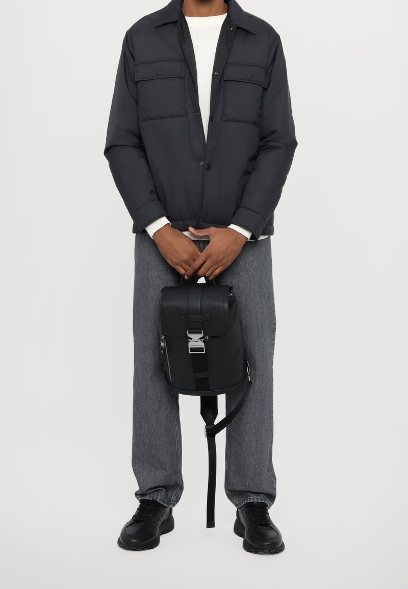 Personne portant une veste noire, un jean gris et des chaussures noires tenant un sac à dos noir avec une poignée supérieure devant son corps.