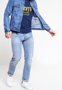 Džínová bunda světle modré barvy, zapínání na knoflíky s kapsami; tmavě modré tričko se žlutým nápisem; světle modré džínsy; bílé tenisky.