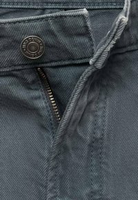 Jeans i en afdæmpet blågrå farve har en lynlås-fly, metal knaplukning og et struktureret stof med et subtilt vævemønster.