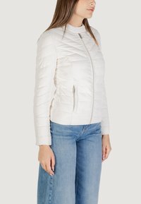 Giacca bianca trapuntata con colletto alto, dotata di chiusura frontale con zip e tasche laterali. Tessuto morbido con una texture liscia.