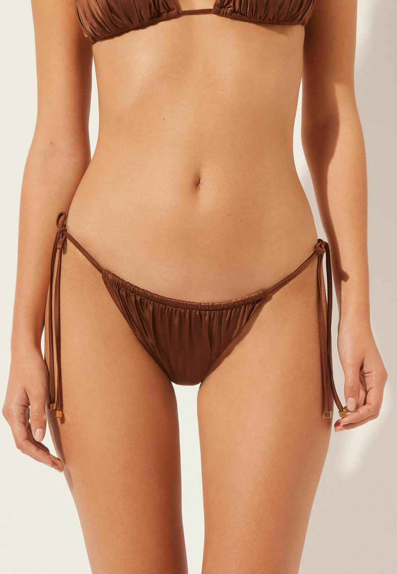 Hnědé bikini kalhotky s nastavitelnými postranními šňůrkami, s nařasenou texturou a minimalistickým designem. Materiál vypadá hladce a jemně.