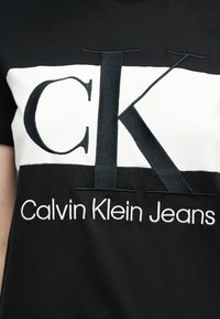Μαύρο και λευκό πουκάμισο με μεγάλο μαύρο κεντητό "CK" και το κείμενο "Calvin Klein Jeans" κάτω ακριβώς στη μέση μπροστά.