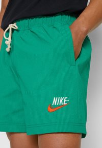 Personne portant un short Nike vert avec un cordon blanc, main dans la poche et logo Nike Swoosh orange visible sur la jambe gauche.