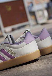 Zapatilla blanca con acentos morados, parte superior de cuero texturizado, diseño de tres franjas y suela de goma. Presenta un detalle del logo en dorado.