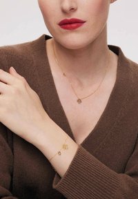 Złoty naszyjnik i bransoletka z motywami kwiatowymi, na tle brązowego swetra. Naszyjnik ma delikatny łańcuszek; bransoletka zawiera podobny urok.