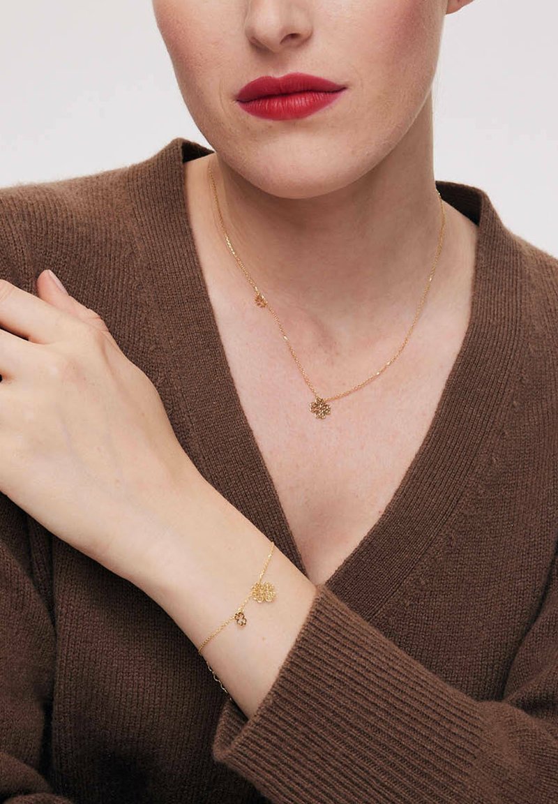 Złoty naszyjnik i bransoletka z motywami kwiatowymi, na tle brązowego swetra. Naszyjnik ma delikatny łańcuszek; bransoletka zawiera podobny urok.